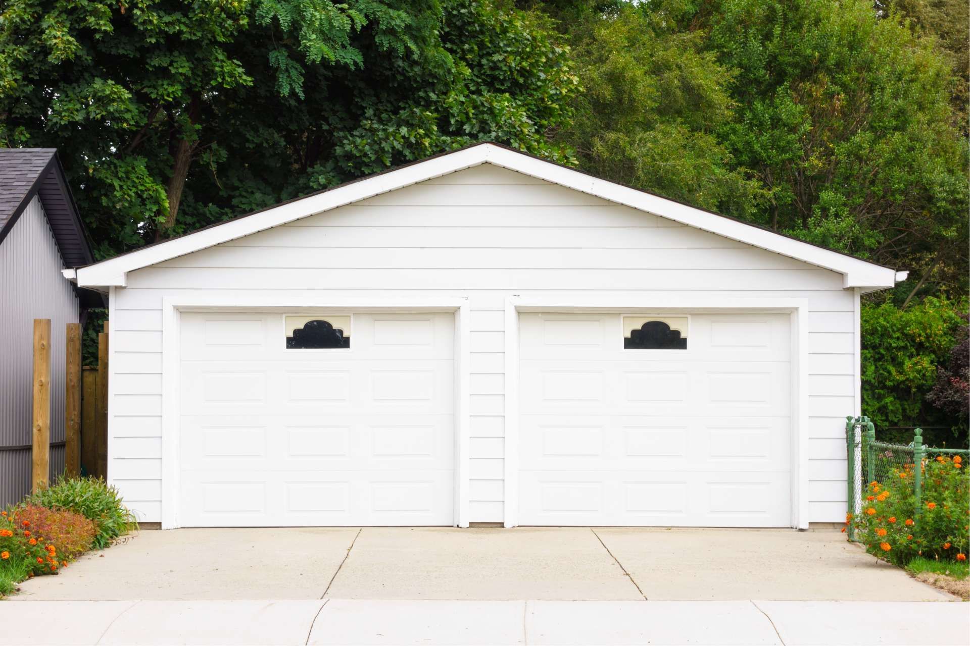 White two-car garage with arched windows, concrete driveway, and foliage backdrop.