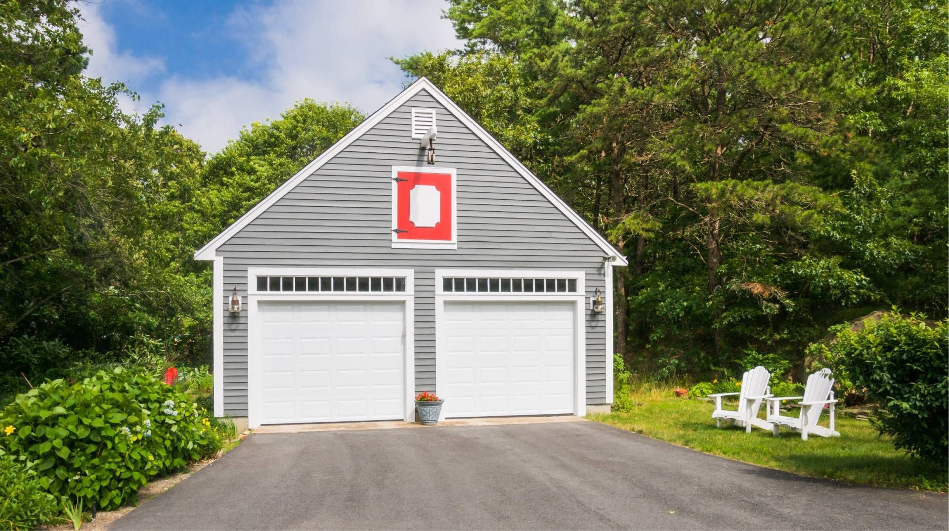 Two-car gray garage with white doors, surrounded by green trees. Two white chairs sit on a green lawn.