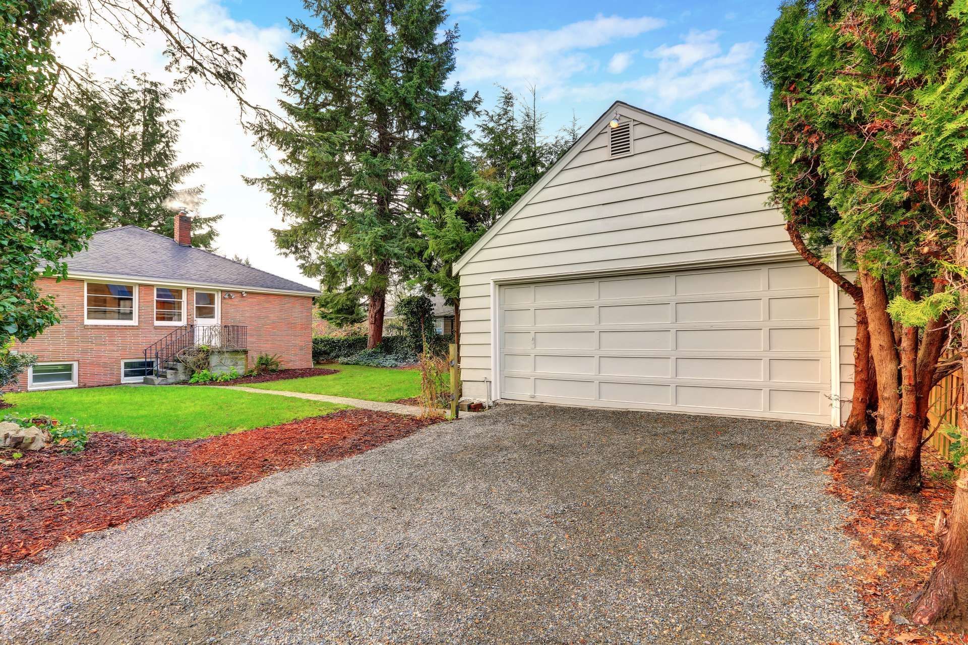 White garage with gravel driveway next to a brick house and evergreen trees.
