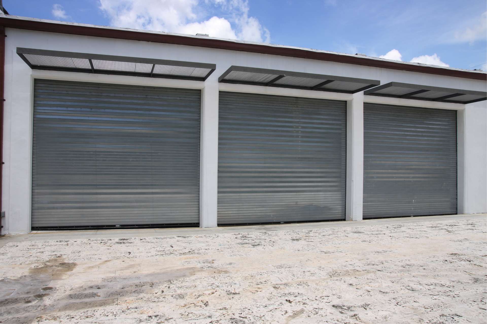 Three gray closed roll-up doors on a white building, under a blue sky.