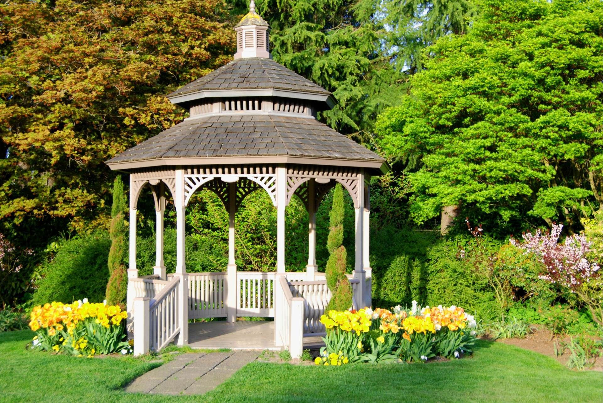 Wooden gazebo in a lush green garden, surrounded by trees and yellow flowers.