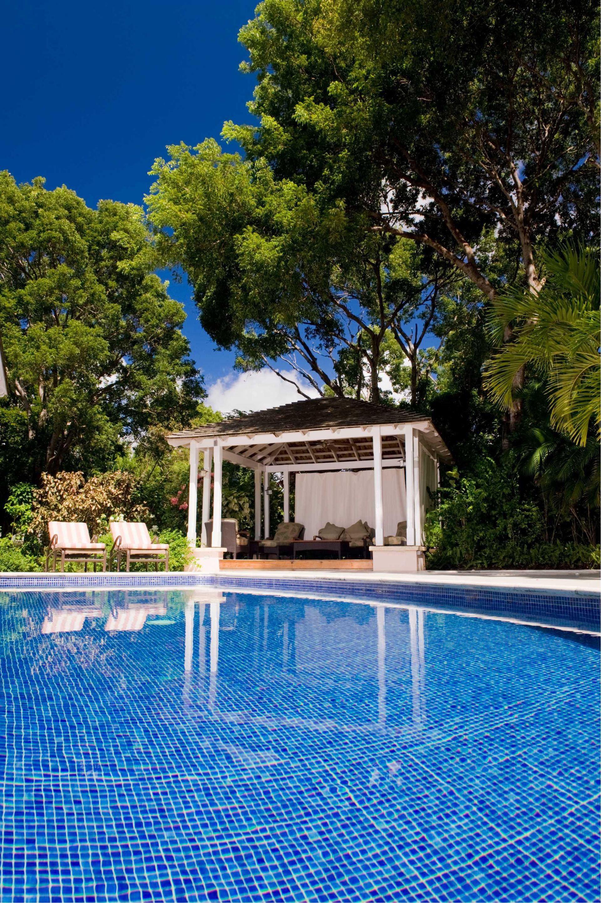 Pool with blue mosaic tiles, white gazebo, lounge chairs, and lush green trees against a blue sky.