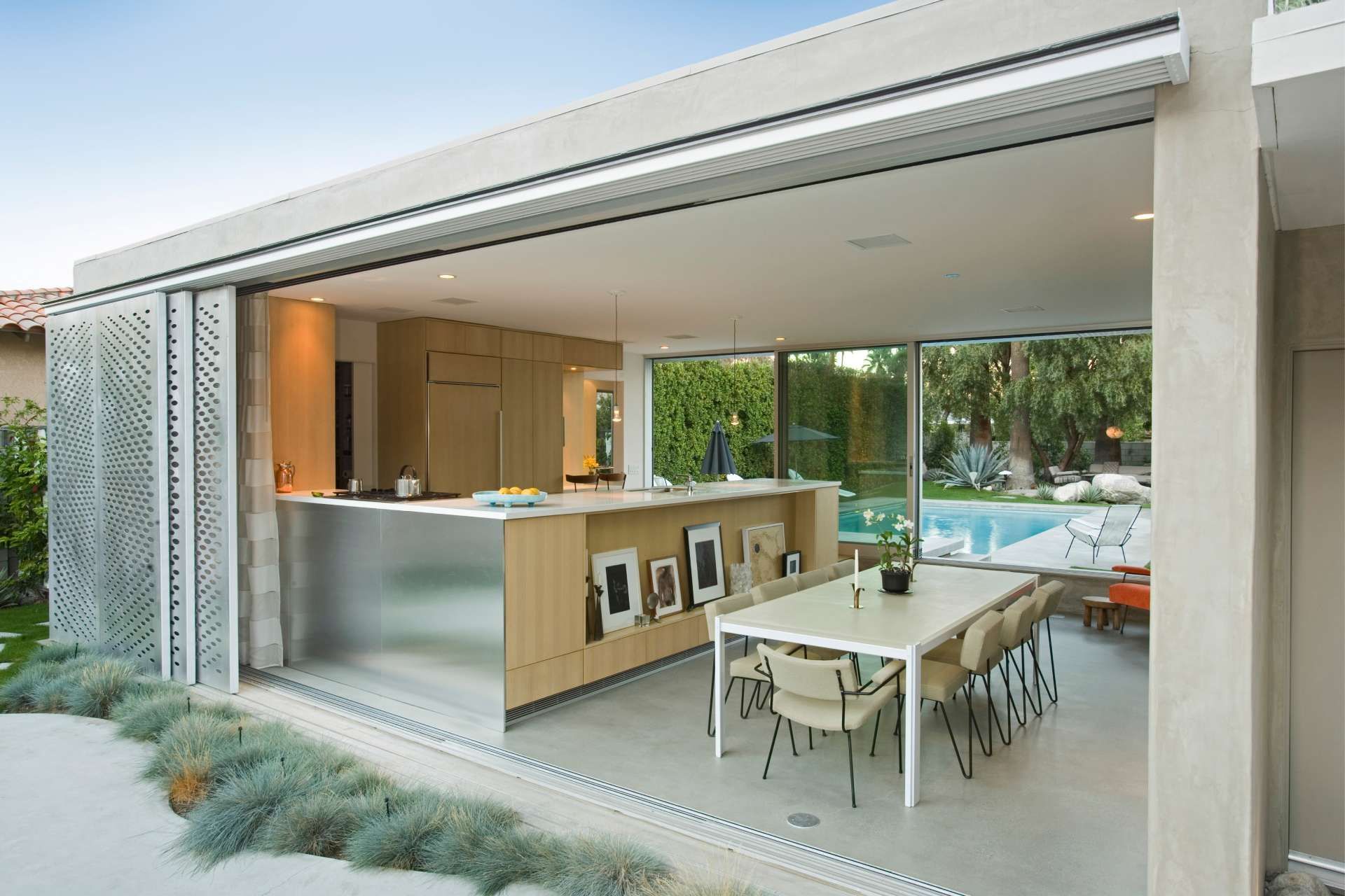 Open-air kitchen with retractable walls, stainless steel island, and dining area overlooking a pool.