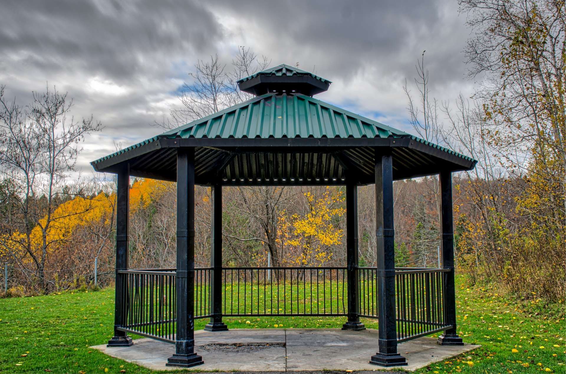 Black gazebo with green roof in a grassy park setting, autumn foliage backdrop, cloudy sky.