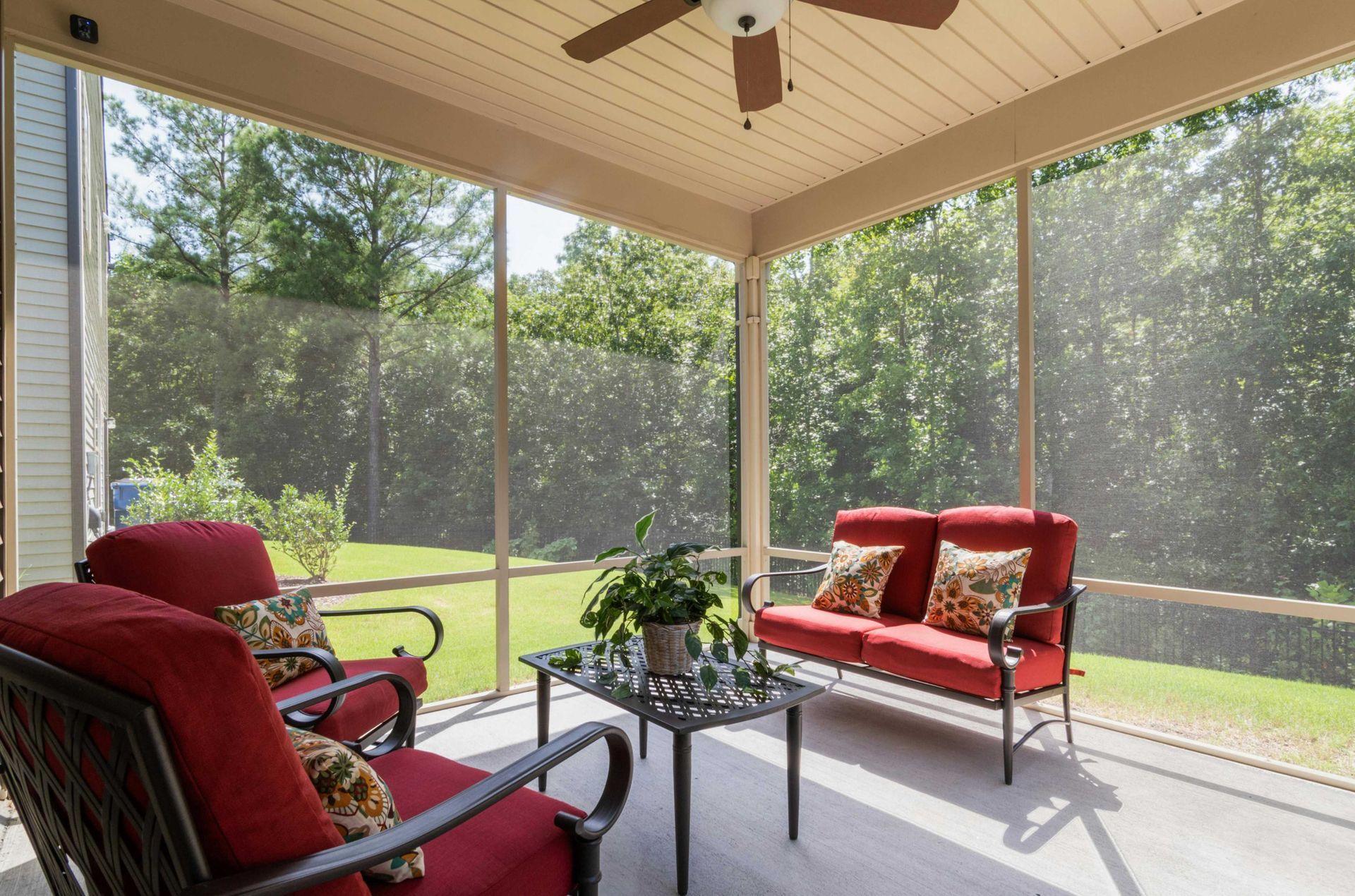 Screened-in porch with red cushioned furniture: a loveseat, two chairs, and a table. Trees and grass in the background.