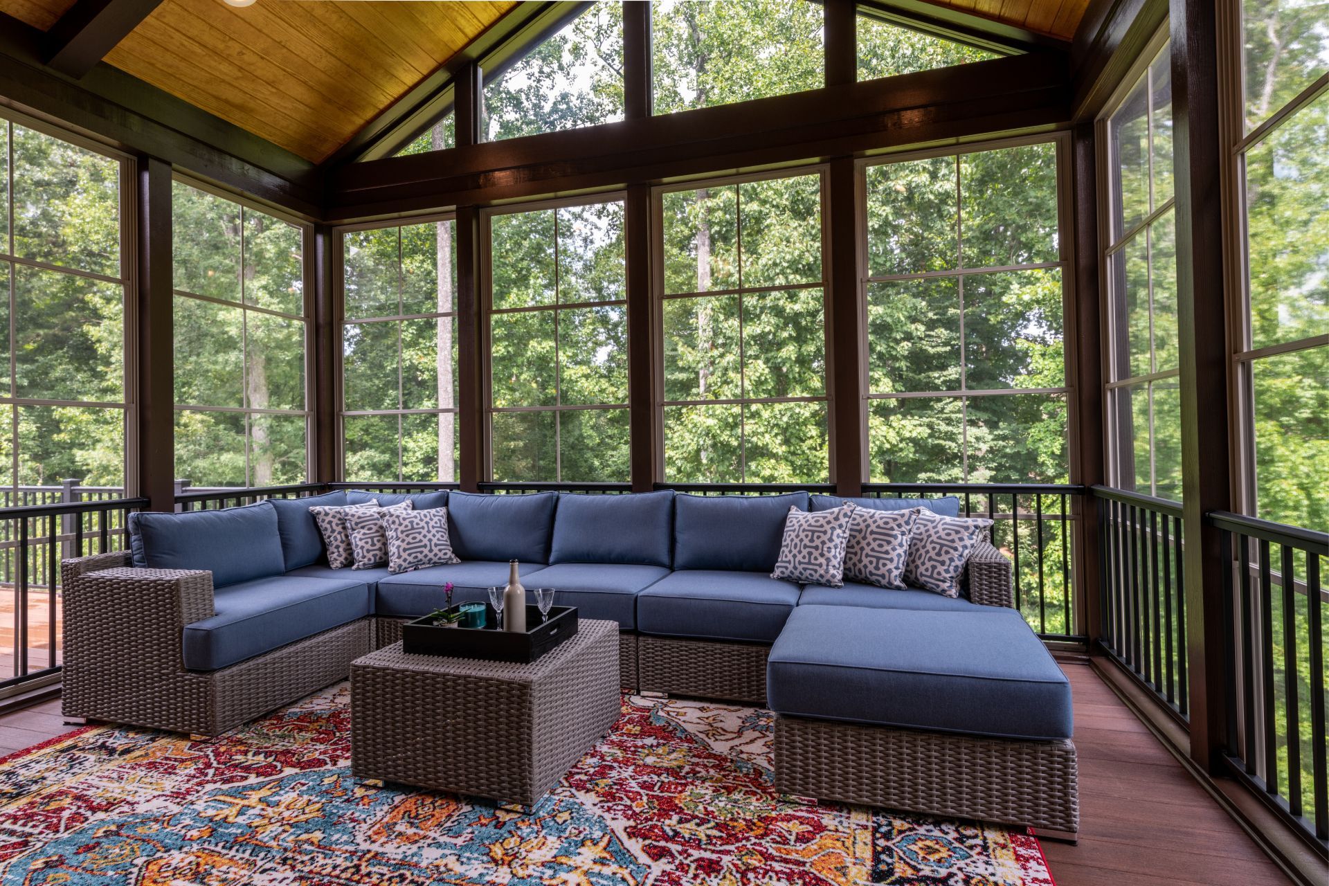 Screened-in porch with blue sectional, patterned rug, and surrounding windows overlooking trees.