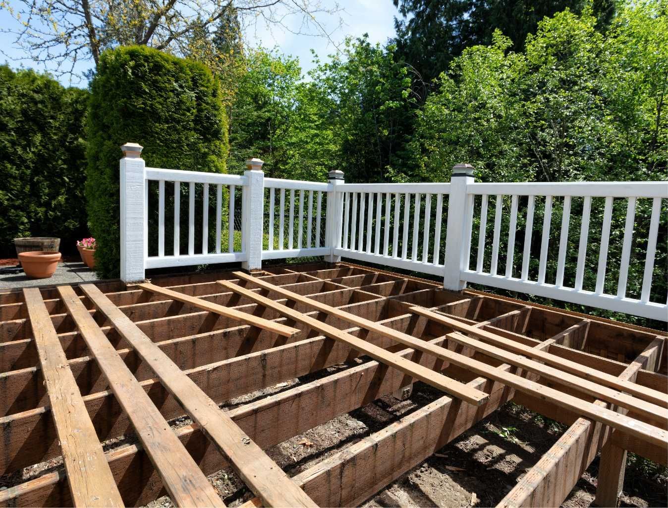 Deck under construction with white railing, wood beams, and surrounding green trees.