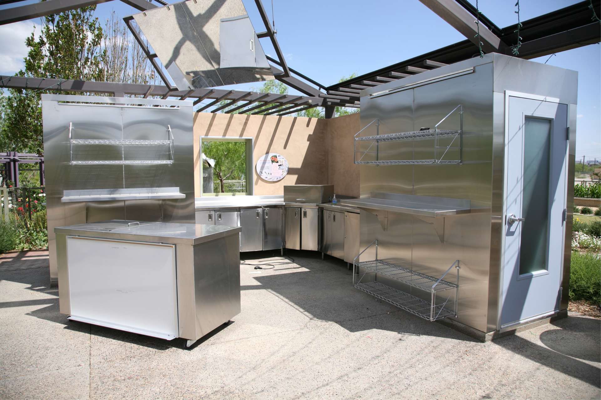 Outdoor stainless steel kitchen with countertops, shelving, and a door, under a wooden trellis.