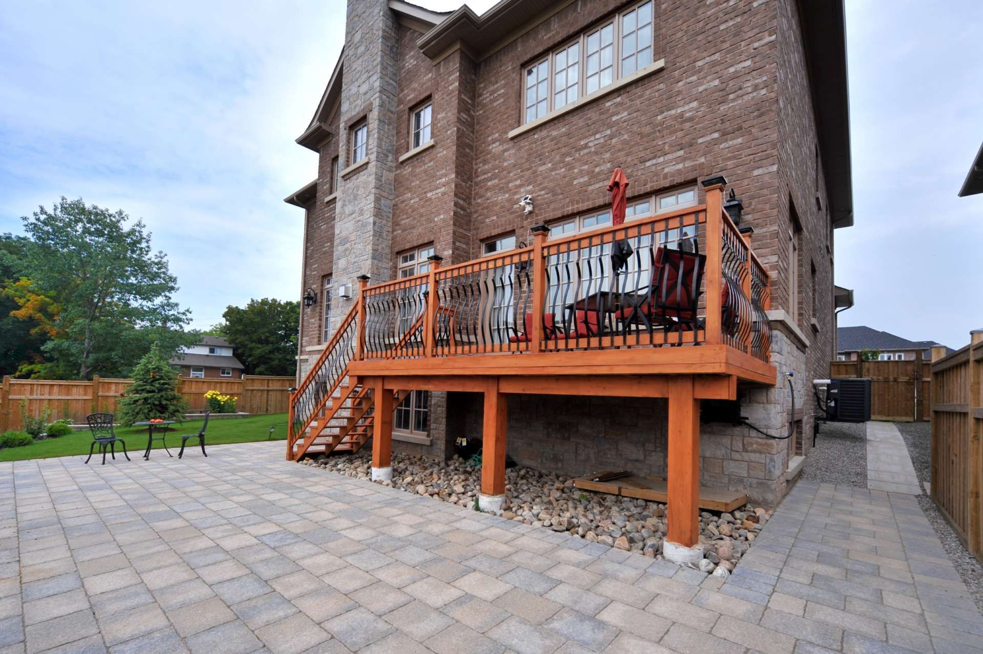 Backyard view of brick house with wooden deck, stone patio, and wooden fence.