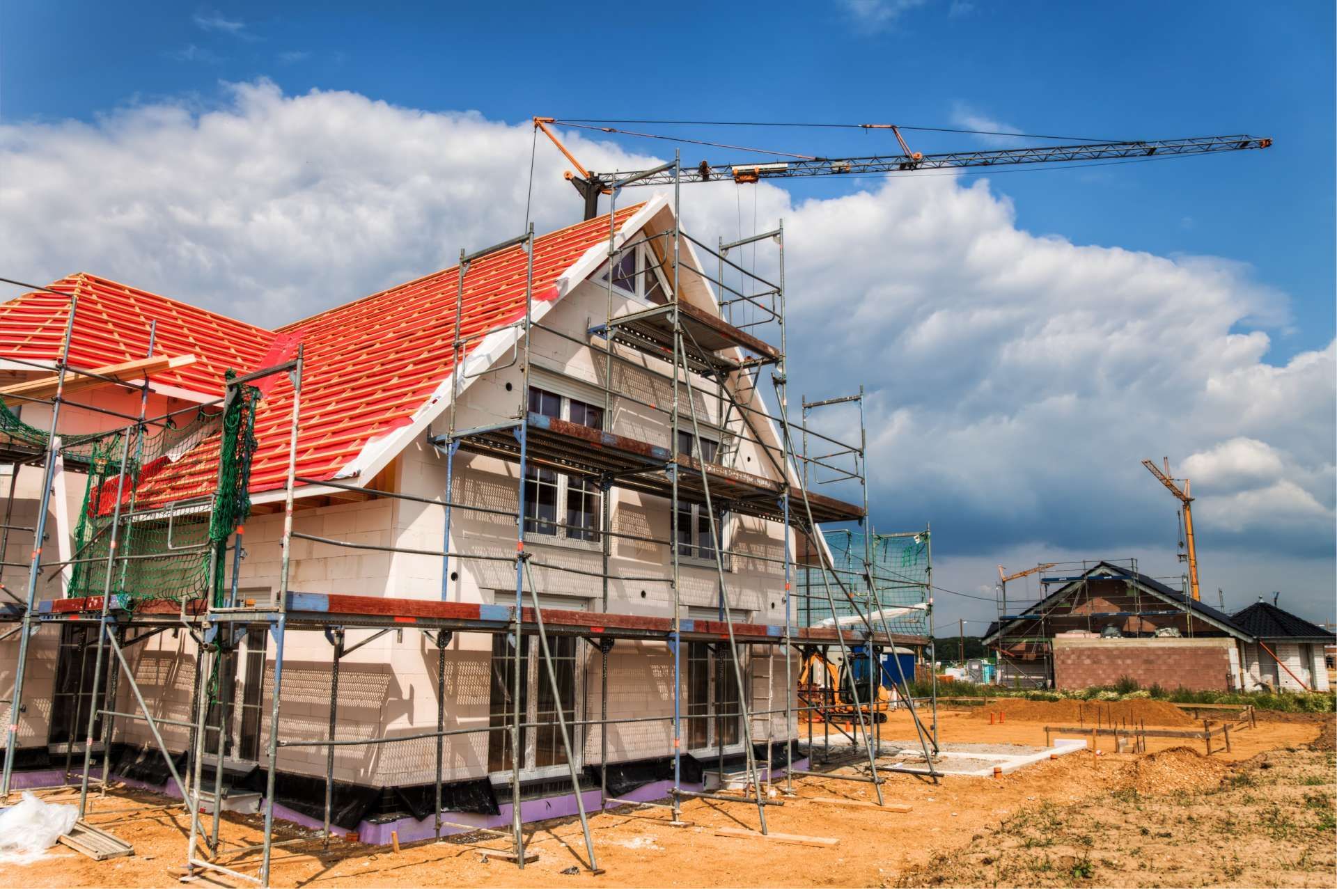 House under construction with scaffolding, red roof, blue sky, and crane.