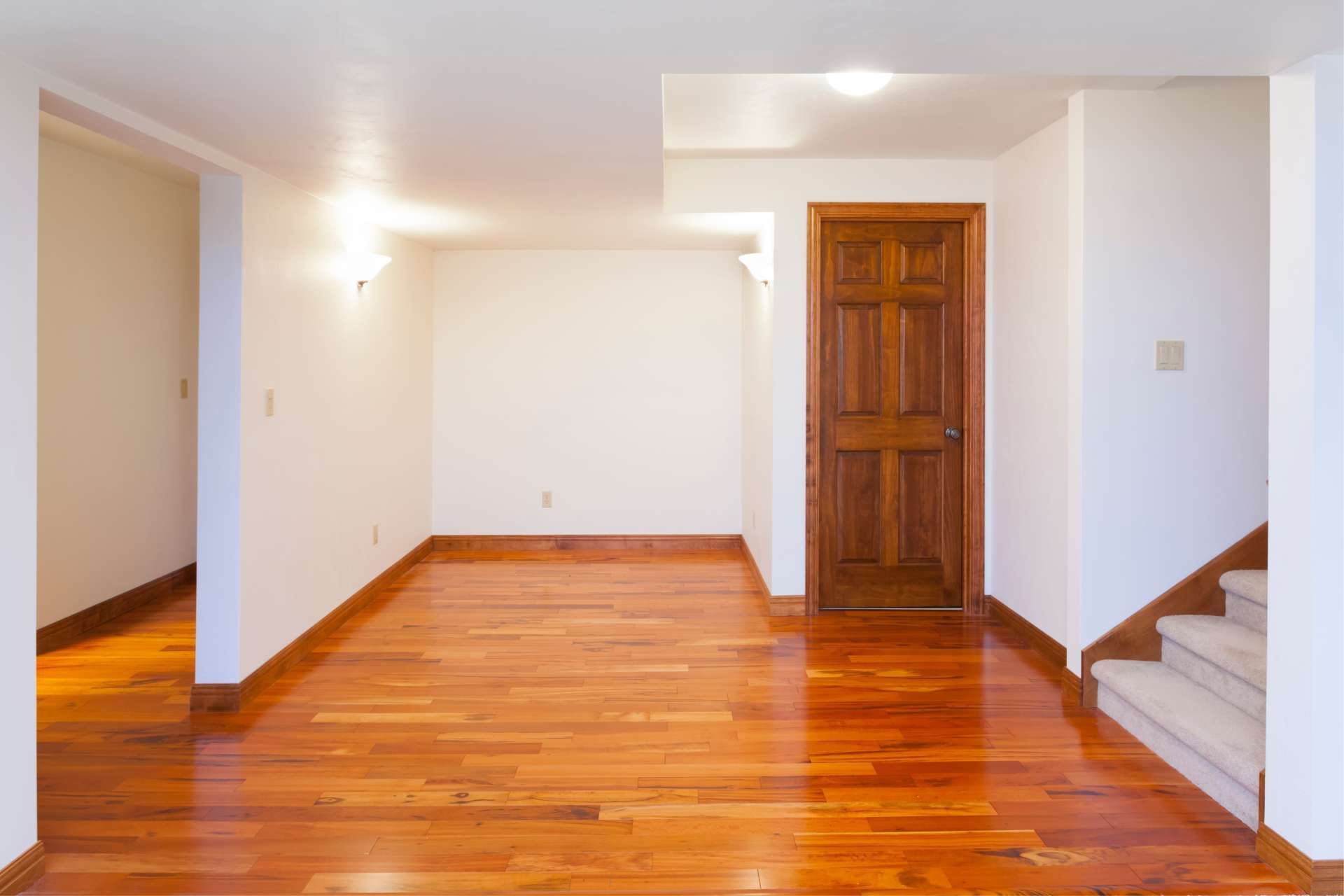 Empty room with hardwood floors, white walls, and a wooden door. Stairs on the right.