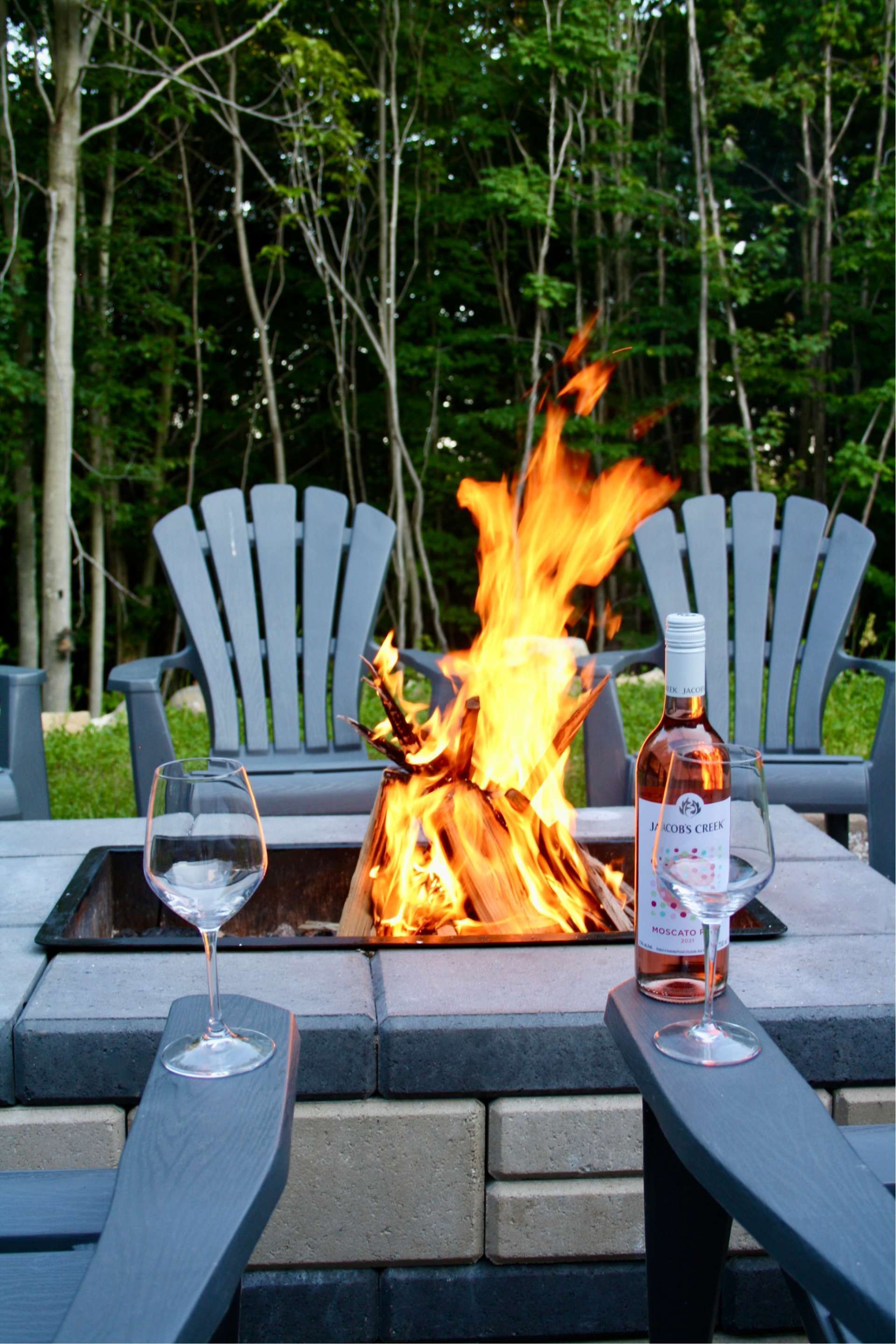 Fire pit with burning flames, wine bottle, and glasses on a patio with two gray chairs, trees in background.