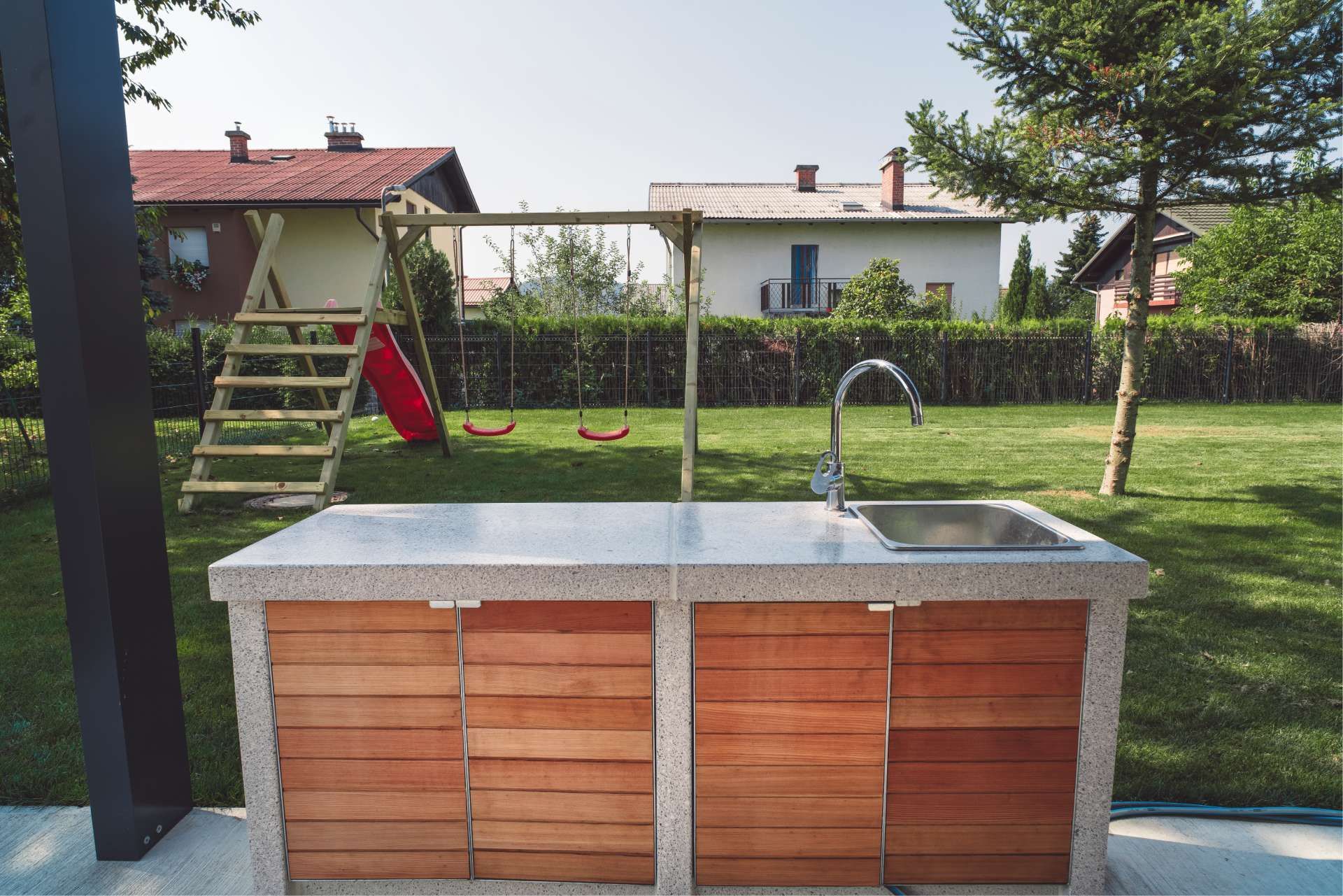 Outdoor kitchen with sink, wooden cabinets, concrete countertop, and a playground in the background.