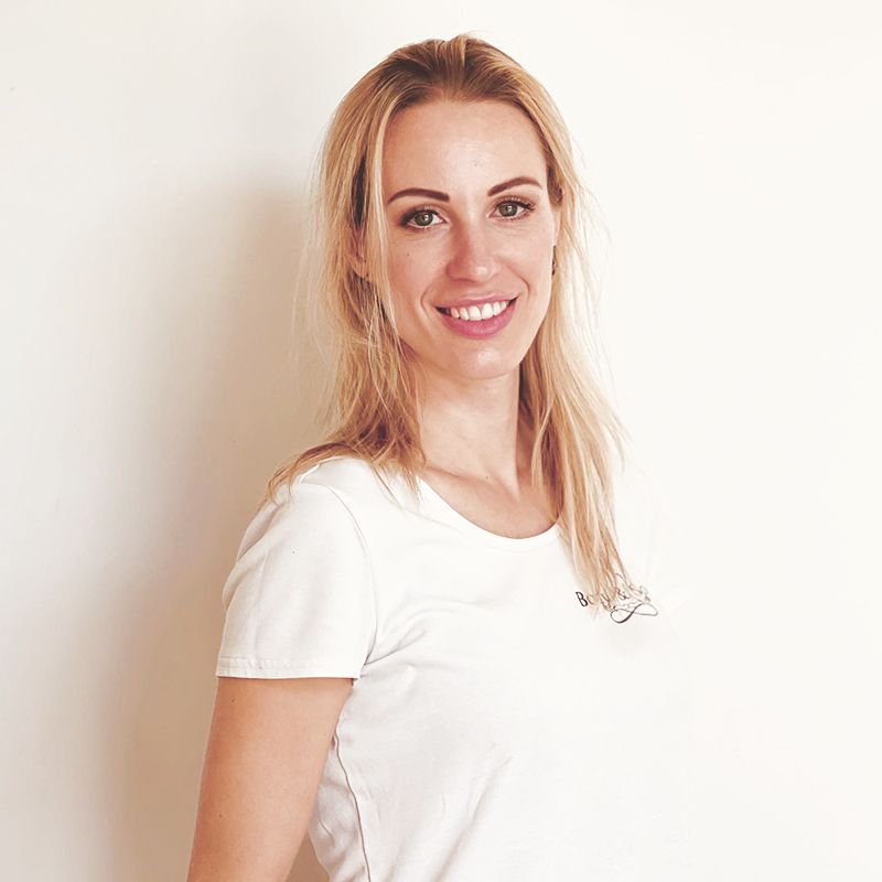 Eine Frau in einem weißen T-Shirt lächelt und steht vor einer weißen Wand.