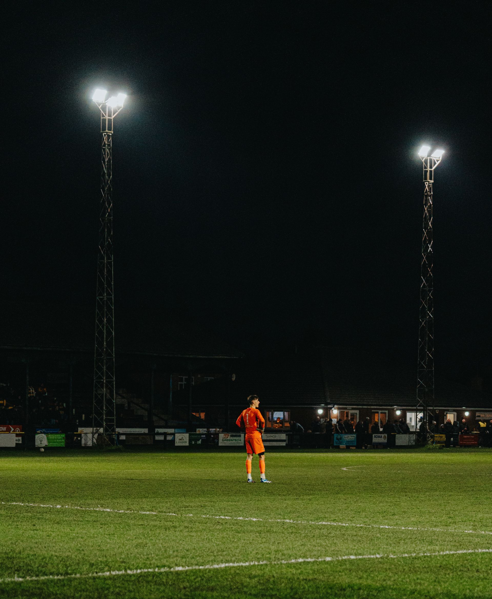 A goalkeeper  is standing on the football pitch at night.