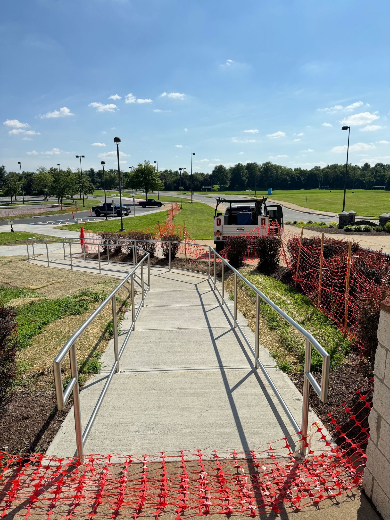 Concrete walkway with metal handrails, leading to a landscaped area, orange safety fencing, blue sky.