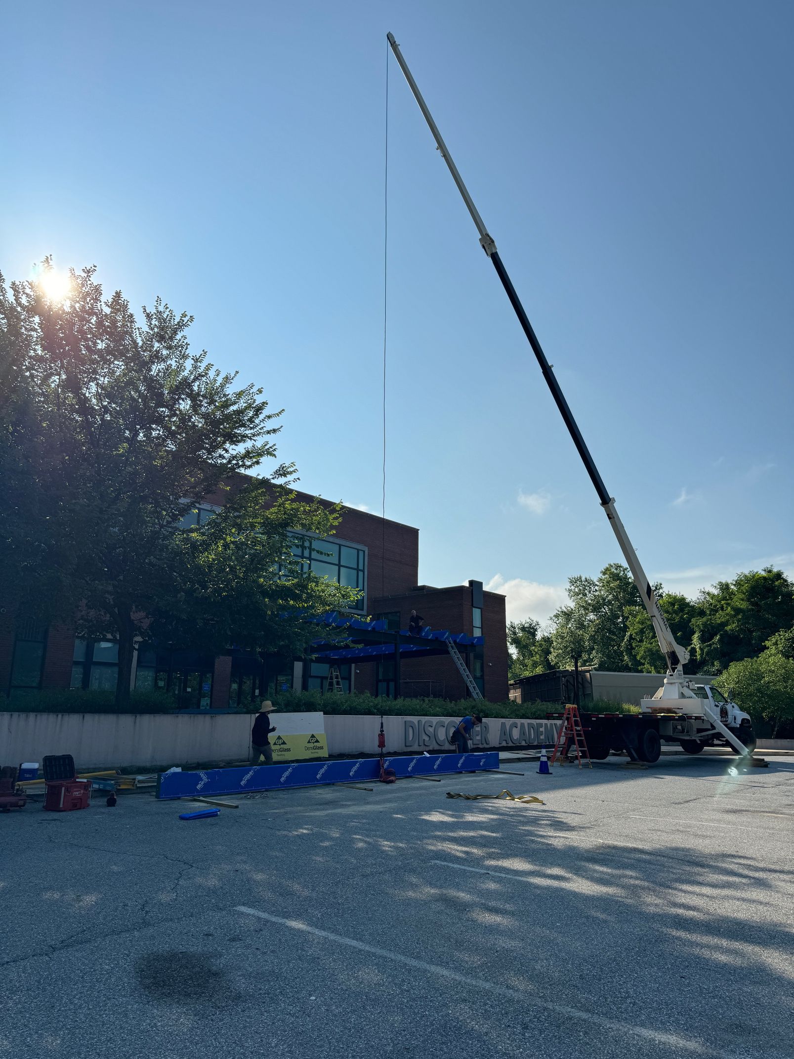 A tall crane is extending upward. Blue safety cones and a building are in the background, under a blue sky.
