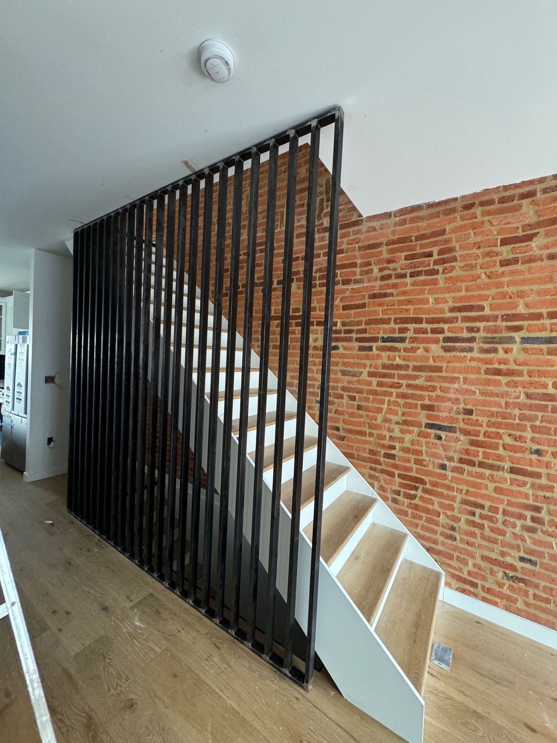 Black vertical railing alongside a staircase with white risers and light-colored treads, set against a brick wall.