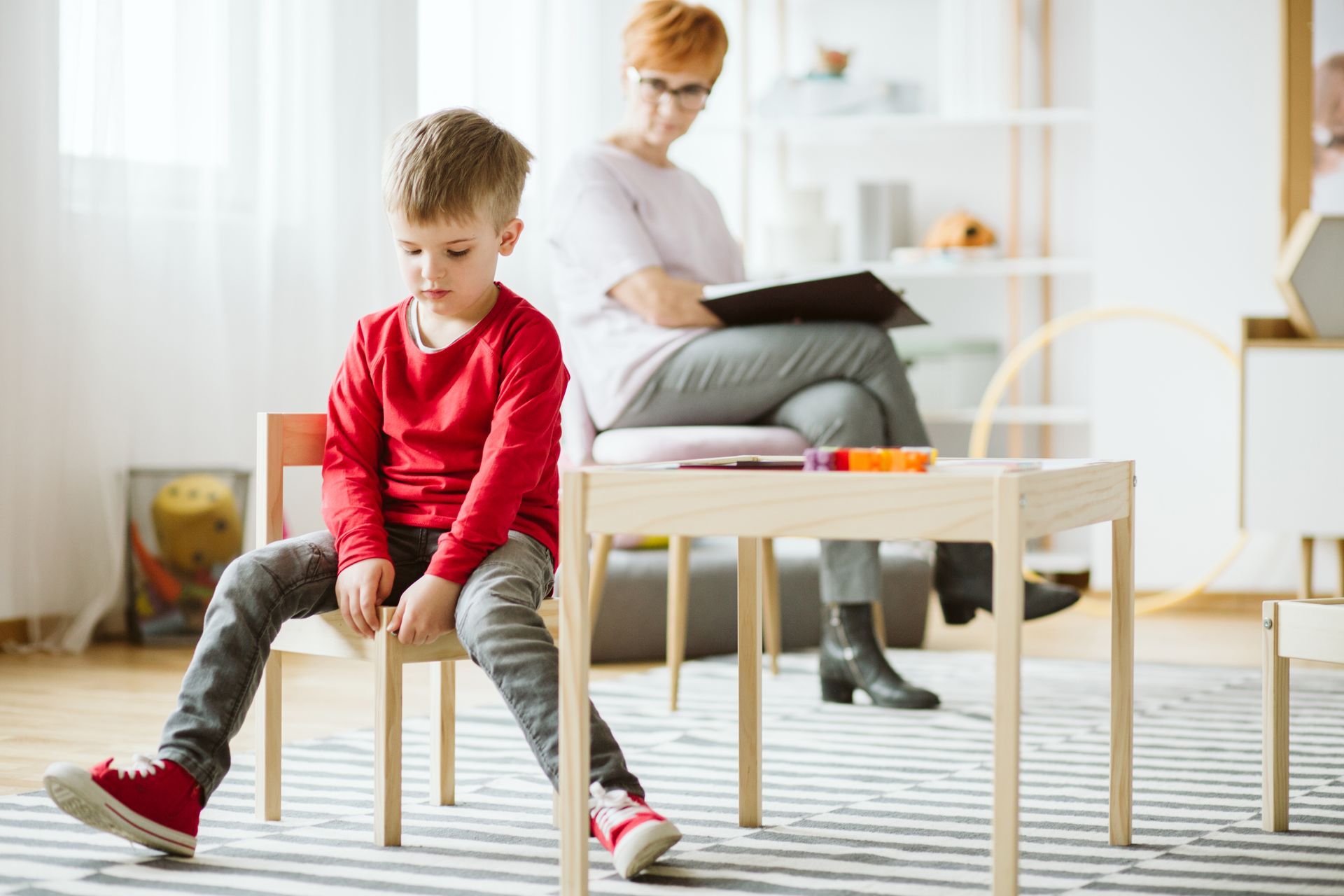 Boy in red shirt sits slumped on chair, at a small table, while a therapist sits and observes.