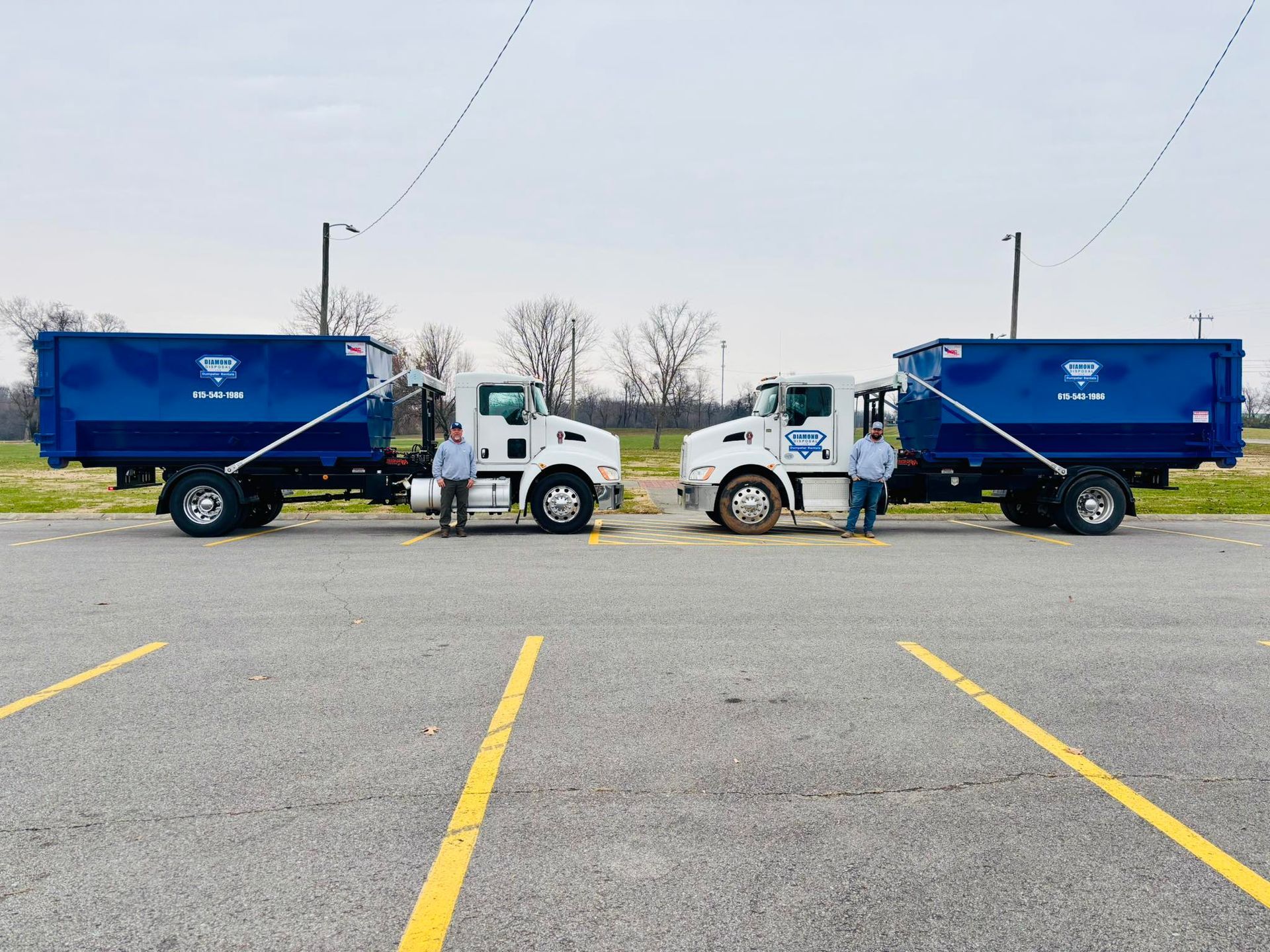 Two white roll-off dump trucks with blue containers parked facing each other with two individuals standing between them.