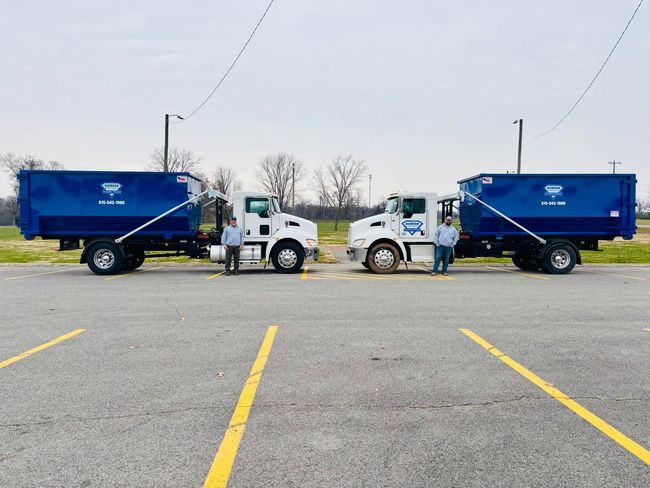 Two commercial dump trucks, one blue and one black, parked side-by-side on an asphalt lot under an overcast sky.