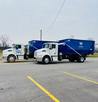 A white dump truck with a large black dumpster container parked on a gravel lot under a cloudy sky.