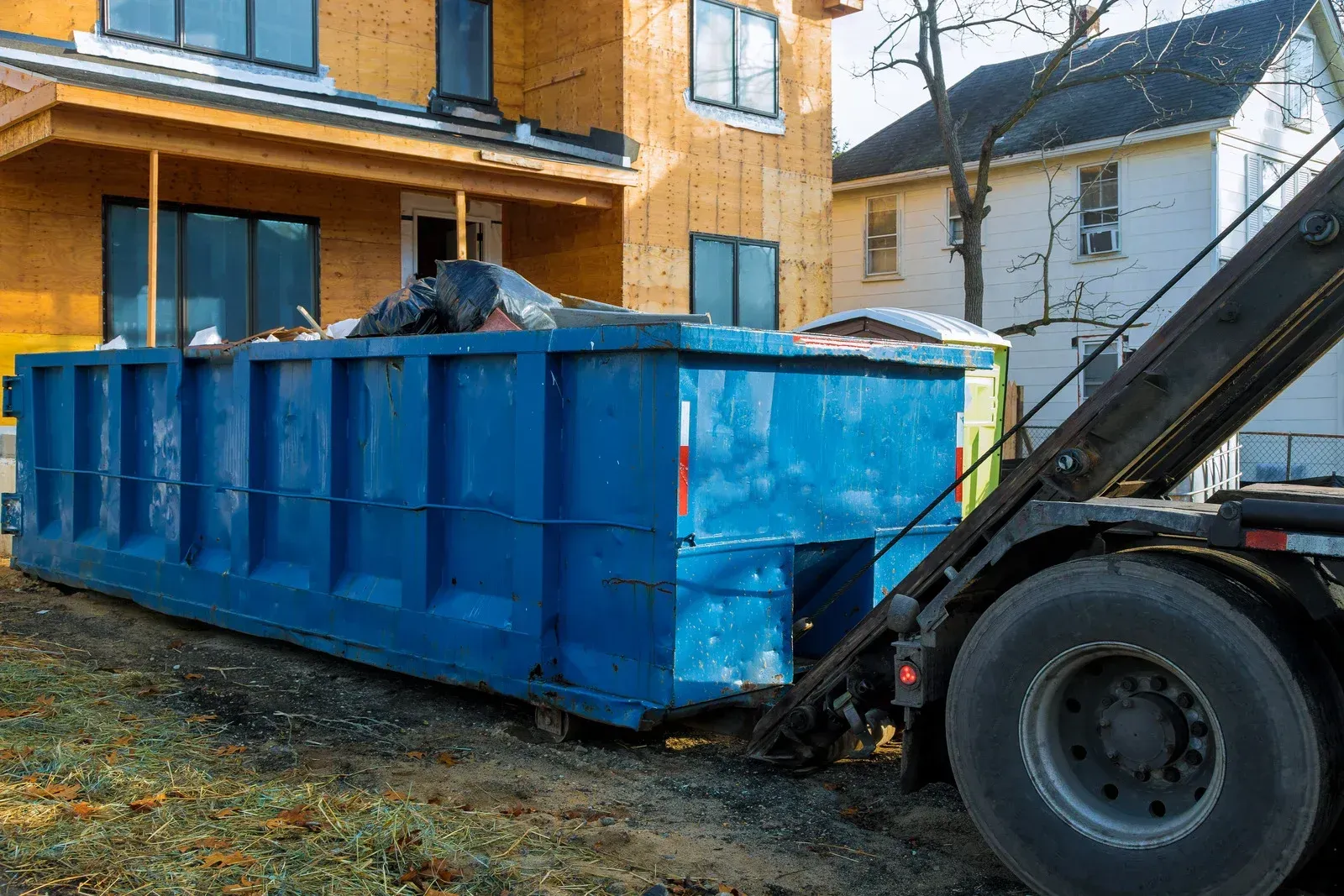 A large blue roll-off dumpster sits on a dirt lot in front of a house under construction.