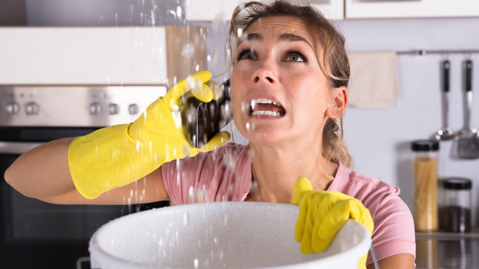 Shocked tenant in yellow gloves catches ceiling leak with a bowl in her kitchen.