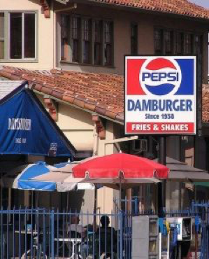 Food establishment, 'Pepsi' & 'Damburger since 1938' signs, outdoor seating under blue/red umbrellas, tiled roof backdrop.