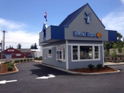 Dutch Bros coffee stand on sunny day, clear blue sky, small blue/white building with company logo, flags atop.