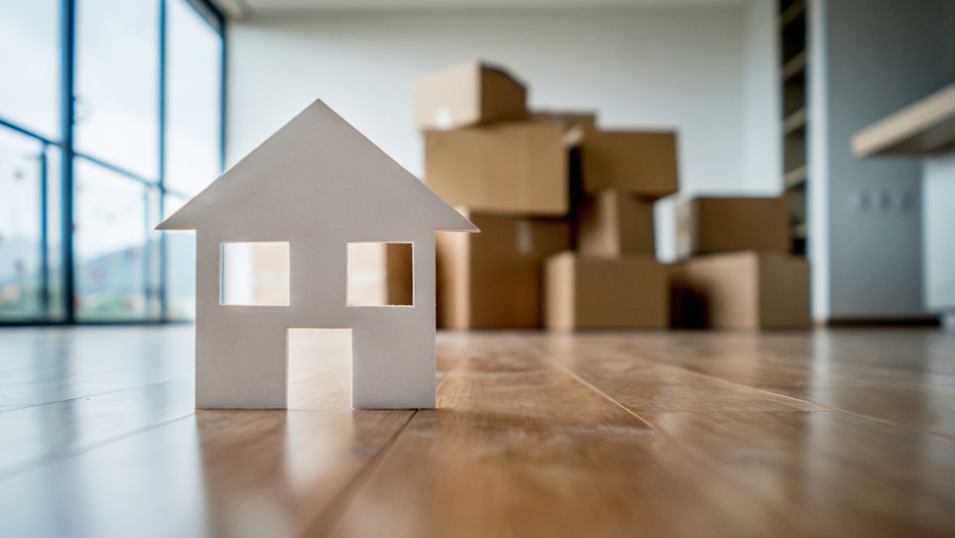 White miniature house on wooden floor with cardboard moving boxes, room with large windows in background.
