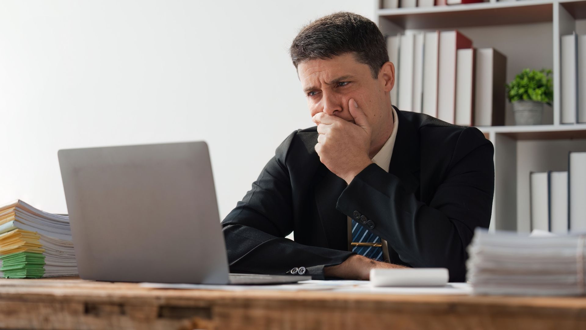 Man in a suit at desk with open laptop, hand on chin, surrounded by documents and folders, shelves in background.