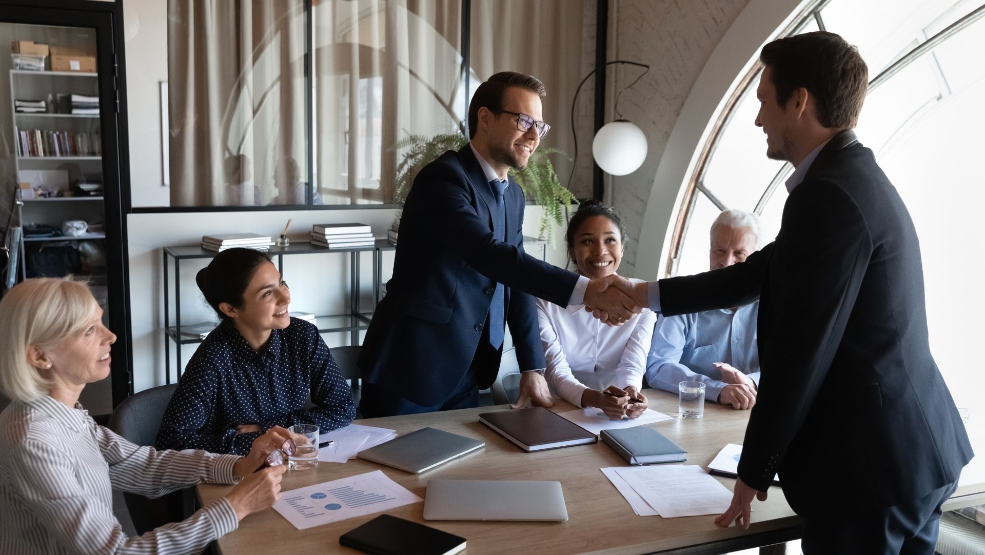 Diverse business professionals in modern office, two men shaking hands, indicating agreement or introduction.