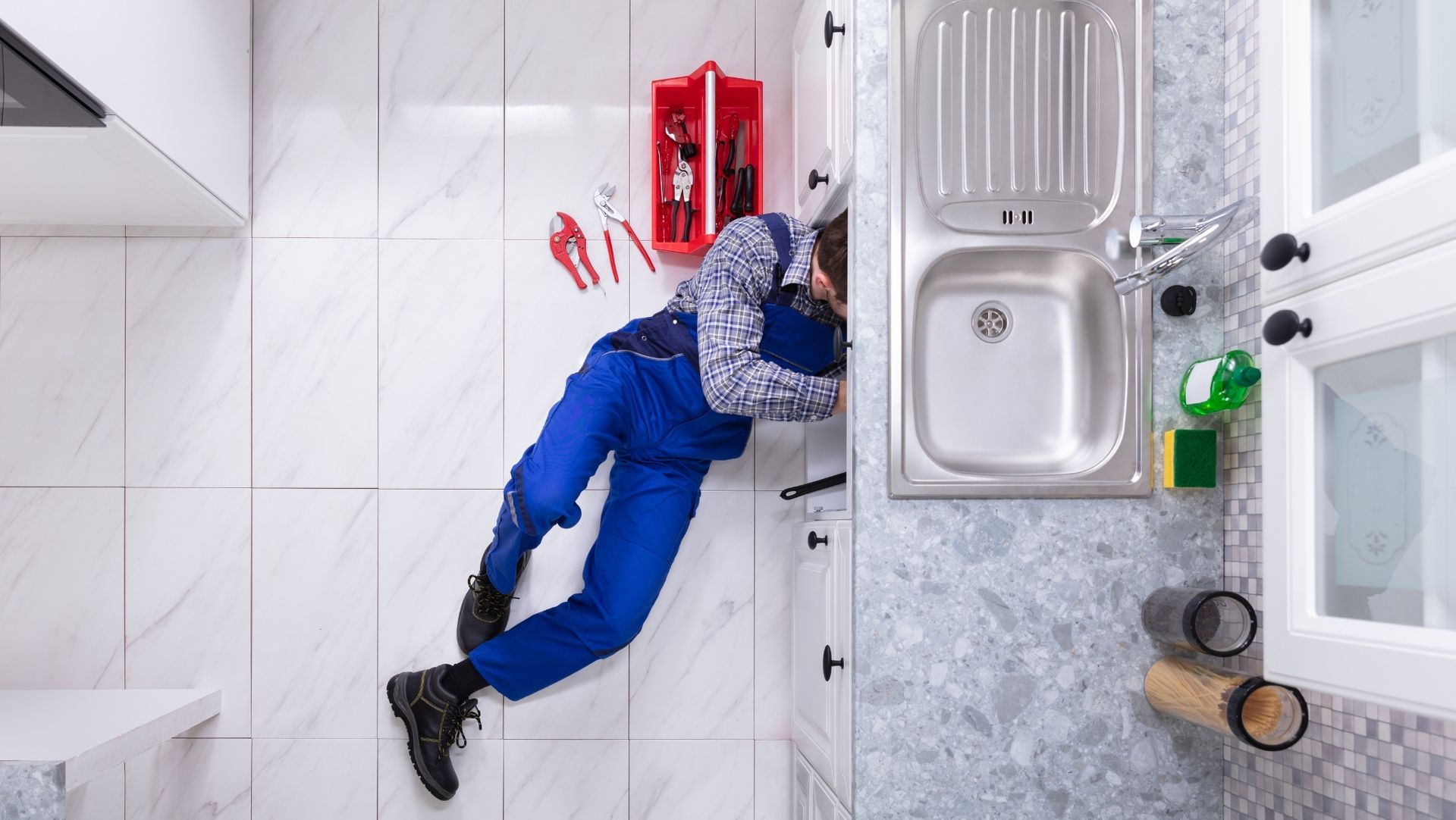 Plumber in blue overalls working under kitchen sink on white tiled floor with red toolbox and scattered tools nearby.
