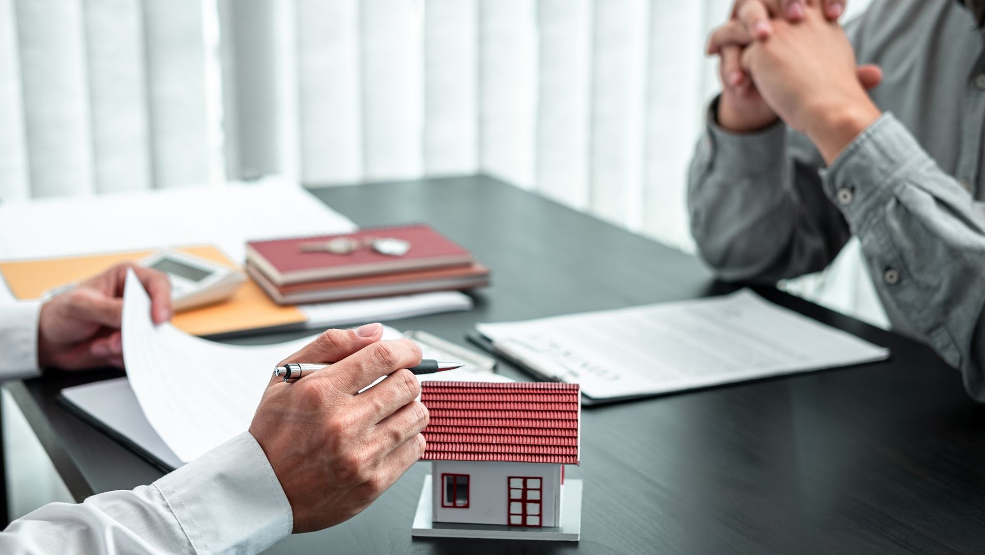 Two people seated at a table with a pen, paper, house model, and documents, discussing real estate or a contract.
