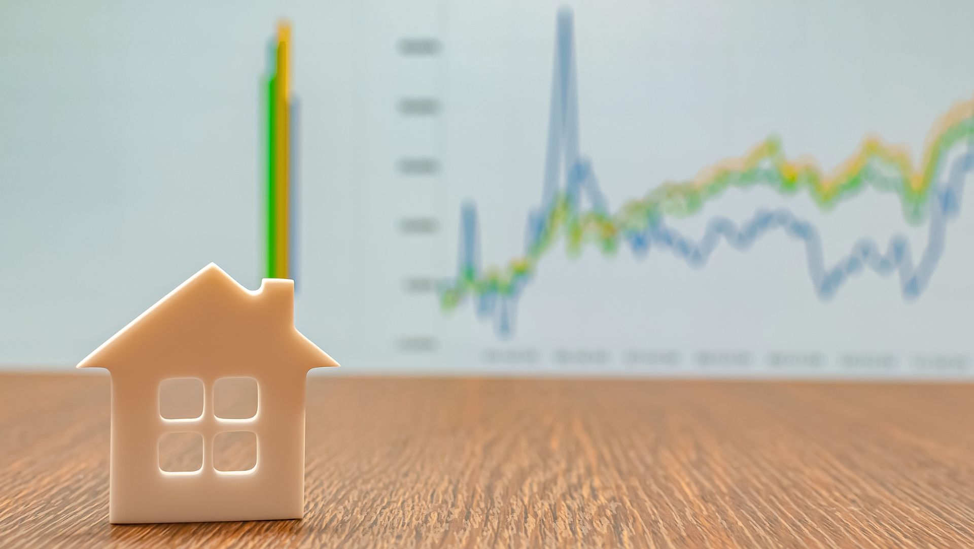 White house model on wooden surface, blurry line graph indicating market trends in the background.