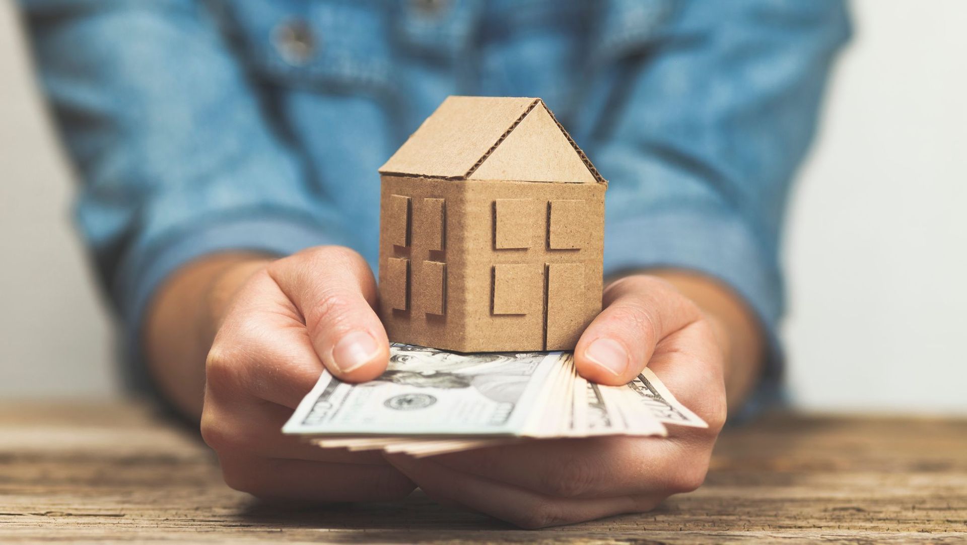 A person holding a small cardboard house model above a stack of money, symbolizing real estate investment or home savings.