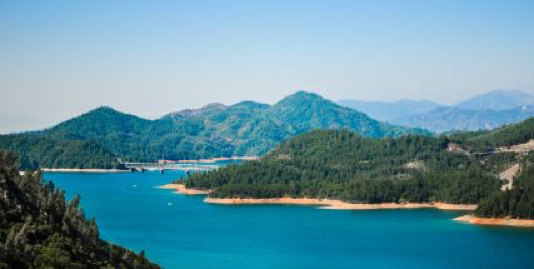 Panoramic view of Shasta Lake, vibrant blue waters surrounded by rolling wooded hills under clear blue sky