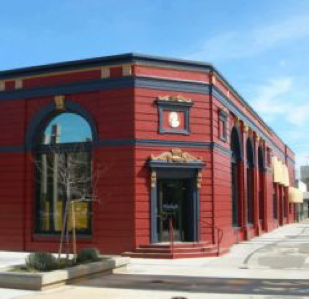 Red and black brick corner building with large windows under a clear blue sky, featuring a small entrance canopy and street-side trees.
