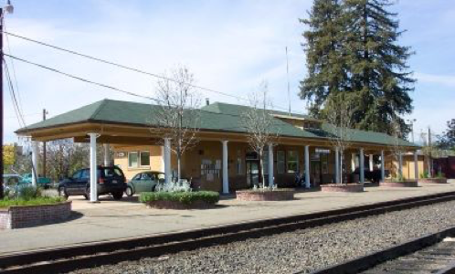 Quaint train station with green roof, tan walls, single railway track, trees, parked cars on sunny day.