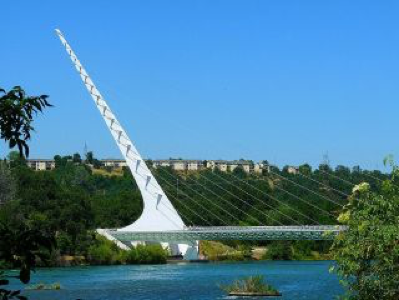 Modern white cable-stayed bridge with tall pylon over tranquil 
river, lush greenery, clear sky. Sundial Bridge, Redding. CA.