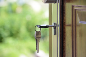 Pair of keys in lock of partially open wooden door, blurred green background visible through doorway.