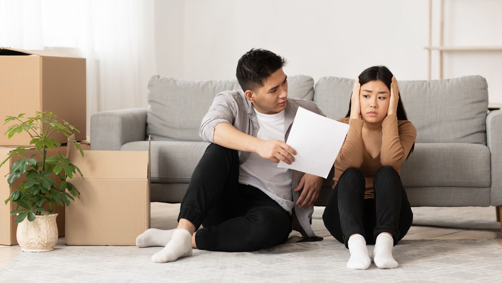 Man and woman sitting on the floor near unpacked moving boxes, looking concerned and talking, indicating a possible move or relocation.