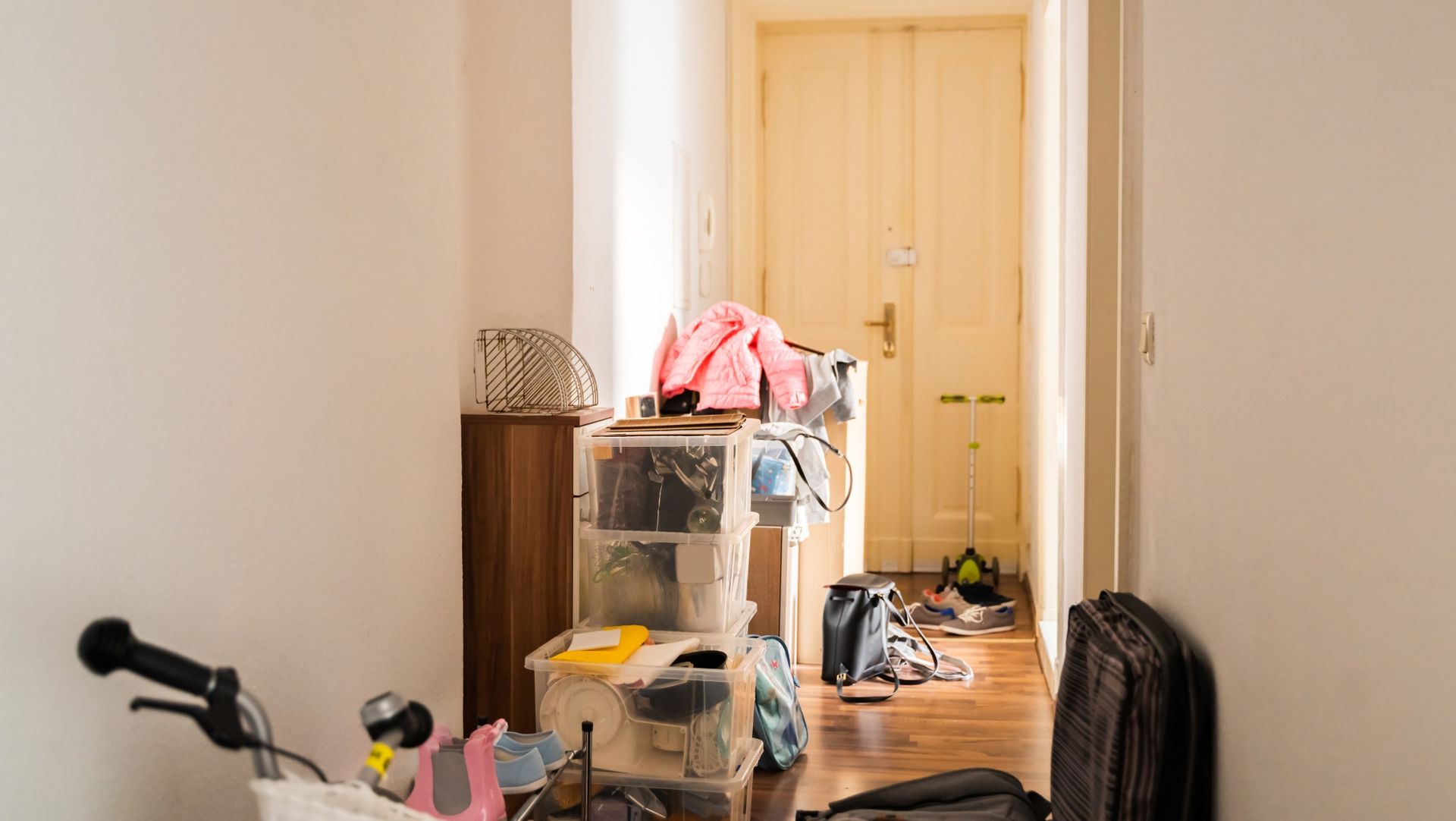 Cluttered hallway with storage bins, bicycle wheel, shoes, bags, pink jacket, and scooter. Wooden floor leads to a closed beige door.
