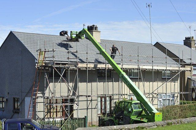 Two workers on scaffolding repair a house roof under a clear blue sky on a sunny day.