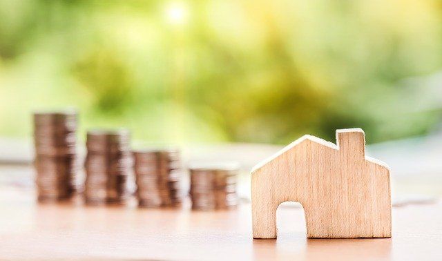 Model of a wooden house with rising coin stacks behind it, symbolizing real estate wealth, against a green blur.