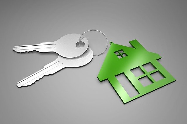 House keys with a green house-shaped keychain on a gray background, symbolizing home ownership or rental.