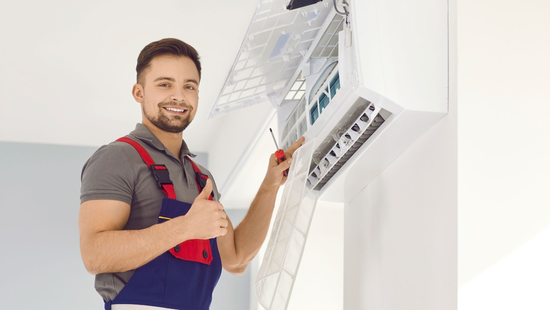 Technician in gray shirt and blue overalls gives thumbs-up while repairing an air conditioning unit.