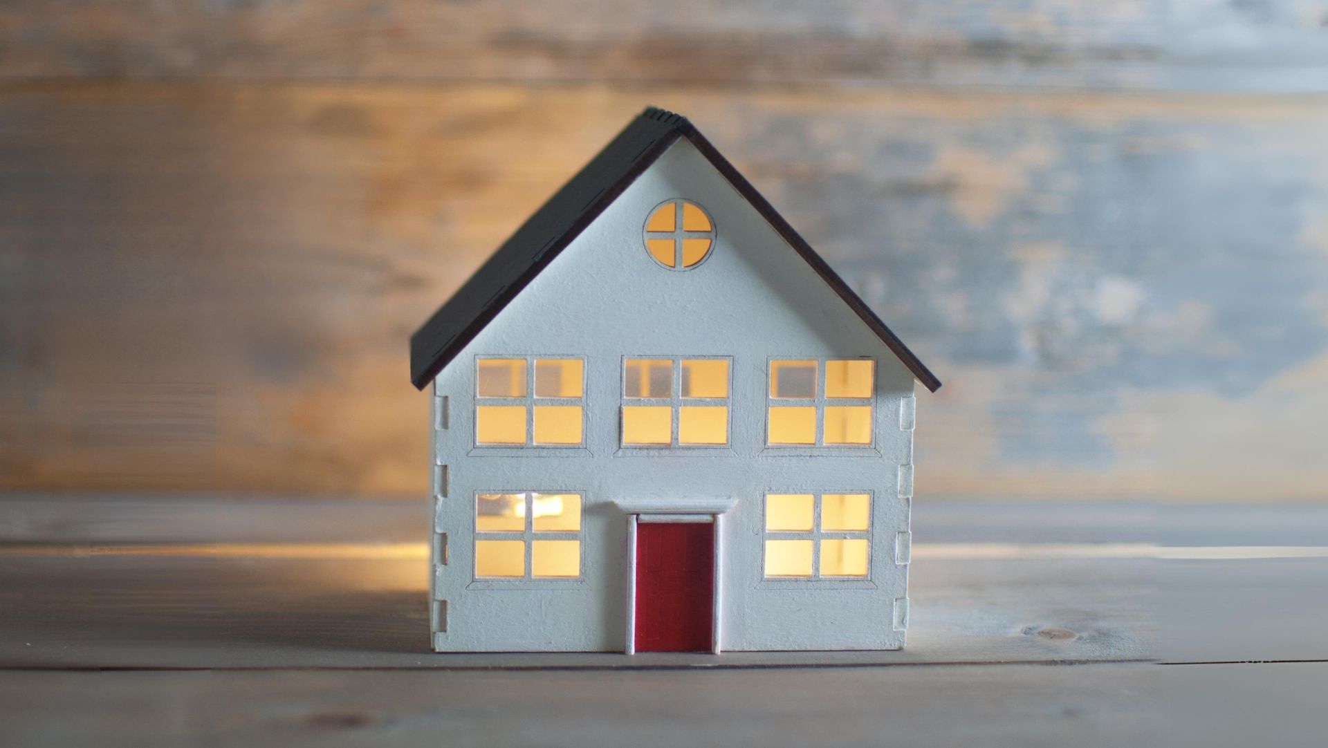 Model house with dark roof, glowing windows, red door on wooden surface, cozy warm lighting.