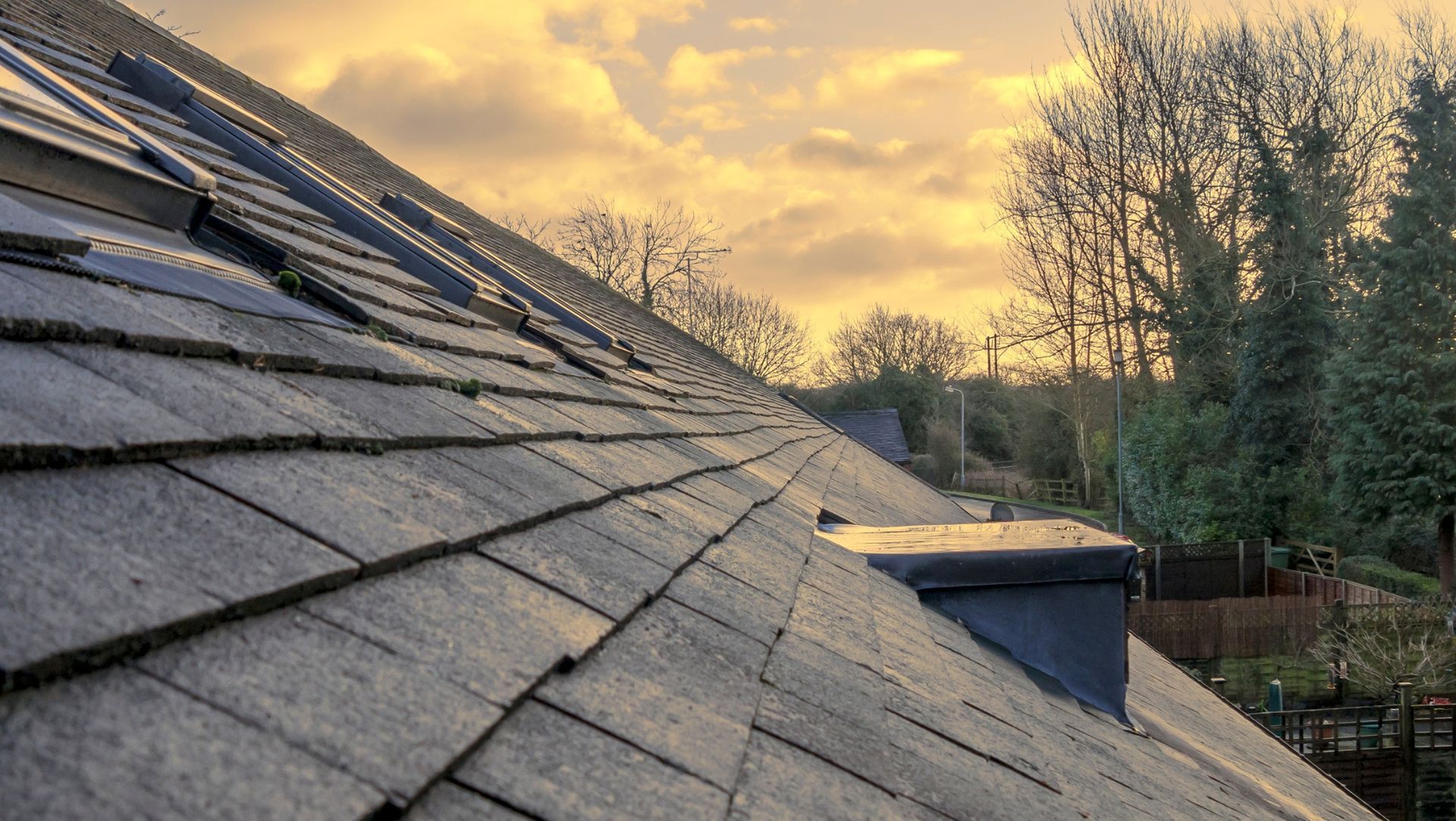 Close-up of sloped slate roof with skylights at sunset, leafless trees and fence in background under golden cloudy sky.